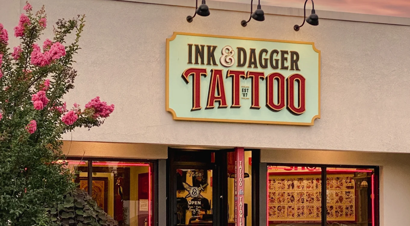 Exterior of the Ink & Dagger Tattoo storefront with the illuminated shop sign above the entrance at dusk